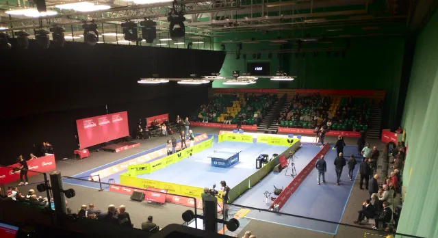 Large sports hall fitted out with black drape, with a single table tennis court in the center, surrounded by green back boards on a blue floor. Lighting rig visible overhead, and a red backdrop podium in the background.